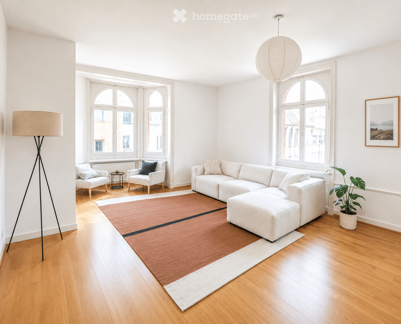 Bright living room with large arched windows, white walls, light wood flooring, a sectional sofa with an ottoman, two armchairs, a small side table, a floor lamp, a potted plant, and a picture frame on the wall.