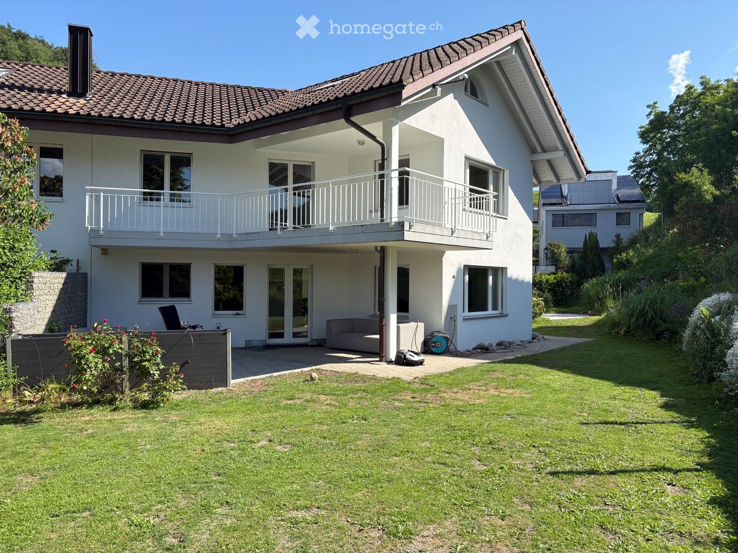 Two-story modern house with white exterior, tiled roof, large windows, balcony, and well-maintained garden.