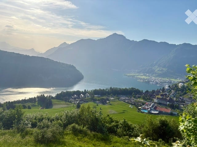 Panoramic view of a lake surrounded by mountains, with a town and green fields in the foreground.