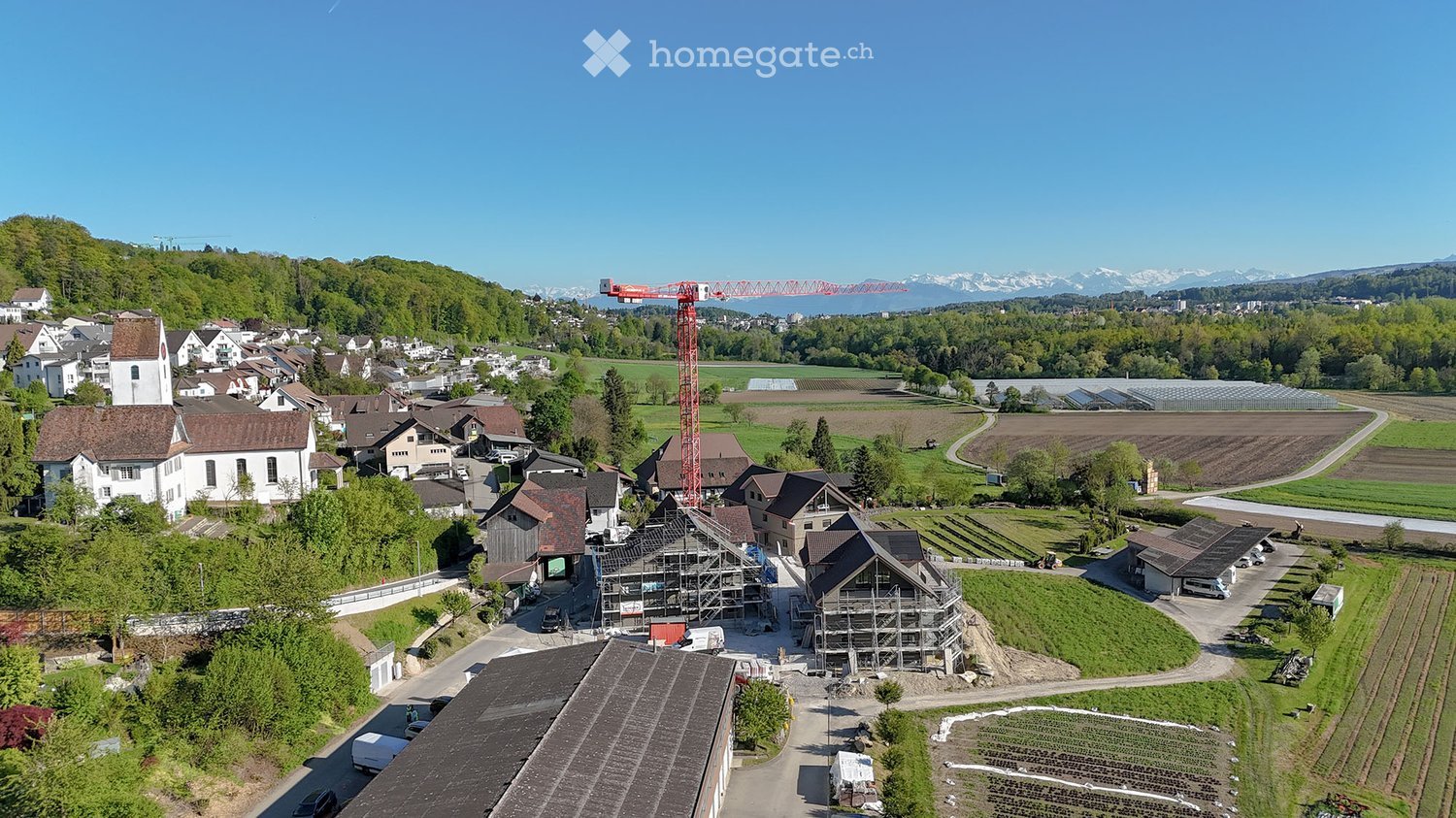 Aerial view of a rural village with traditional houses, a church, and a construction site with a crane in the background, surrounded by green fields and mountains.
