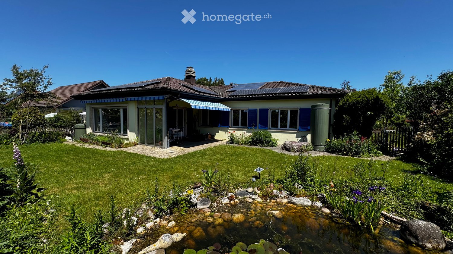 Two-story house with solar panels on the roof, blue shutters, and a well-maintained garden with a pond and patio area.