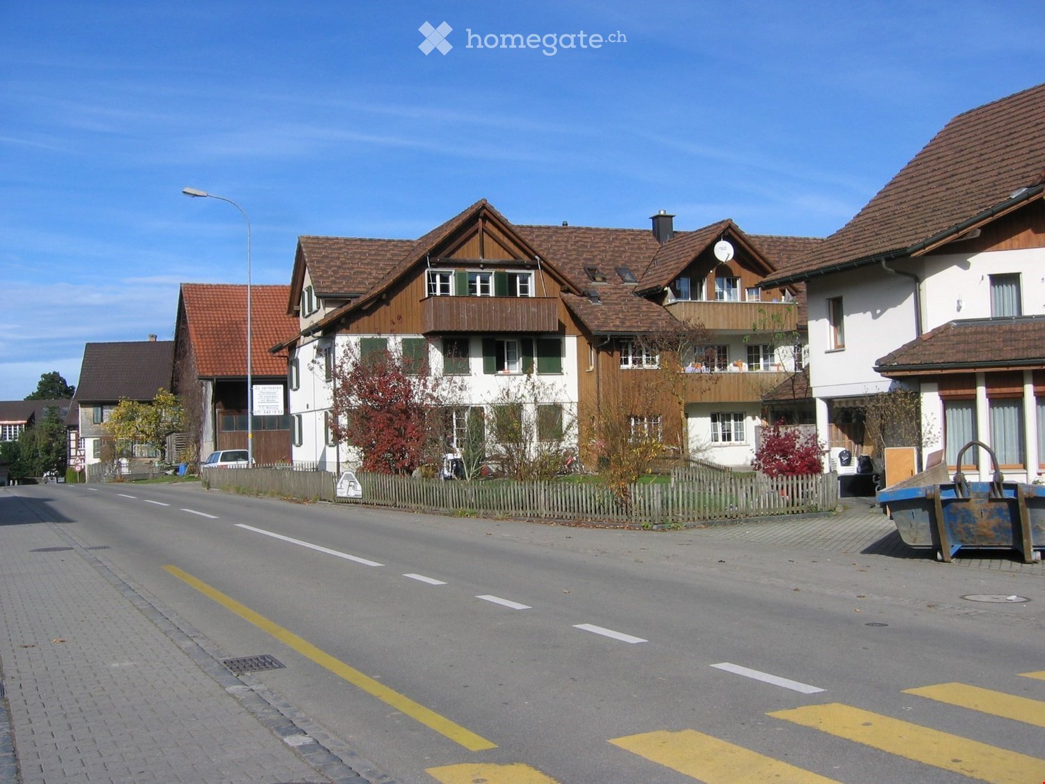 Residential street with traditional Swiss houses featuring wooden facades, tiled roofs, and balconies. The road is paved with clear markings, and there are small fences and trees lining the street.