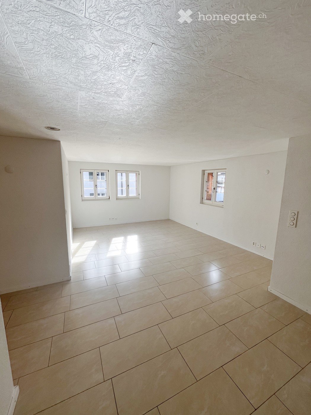 Empty room with light-colored tiled flooring and white walls, featuring three windows and a textured ceiling.