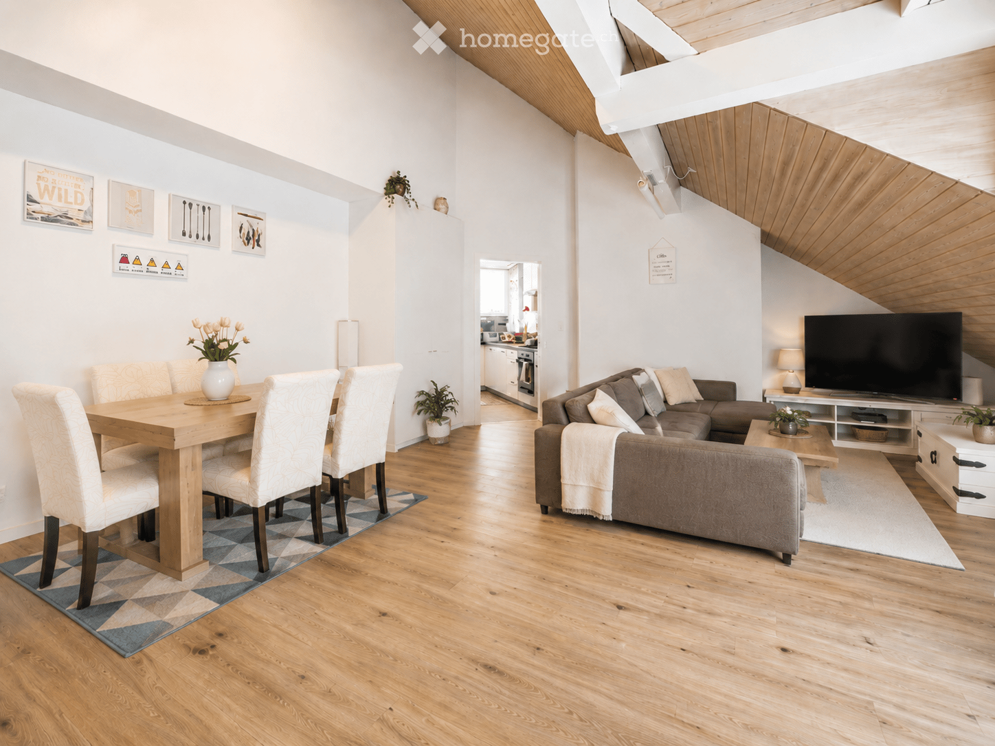 Open-plan living and dining area with wooden flooring, a grey sectional sofa, a wooden dining table with four chairs, a television on a stand, and decorative vases with flowers.