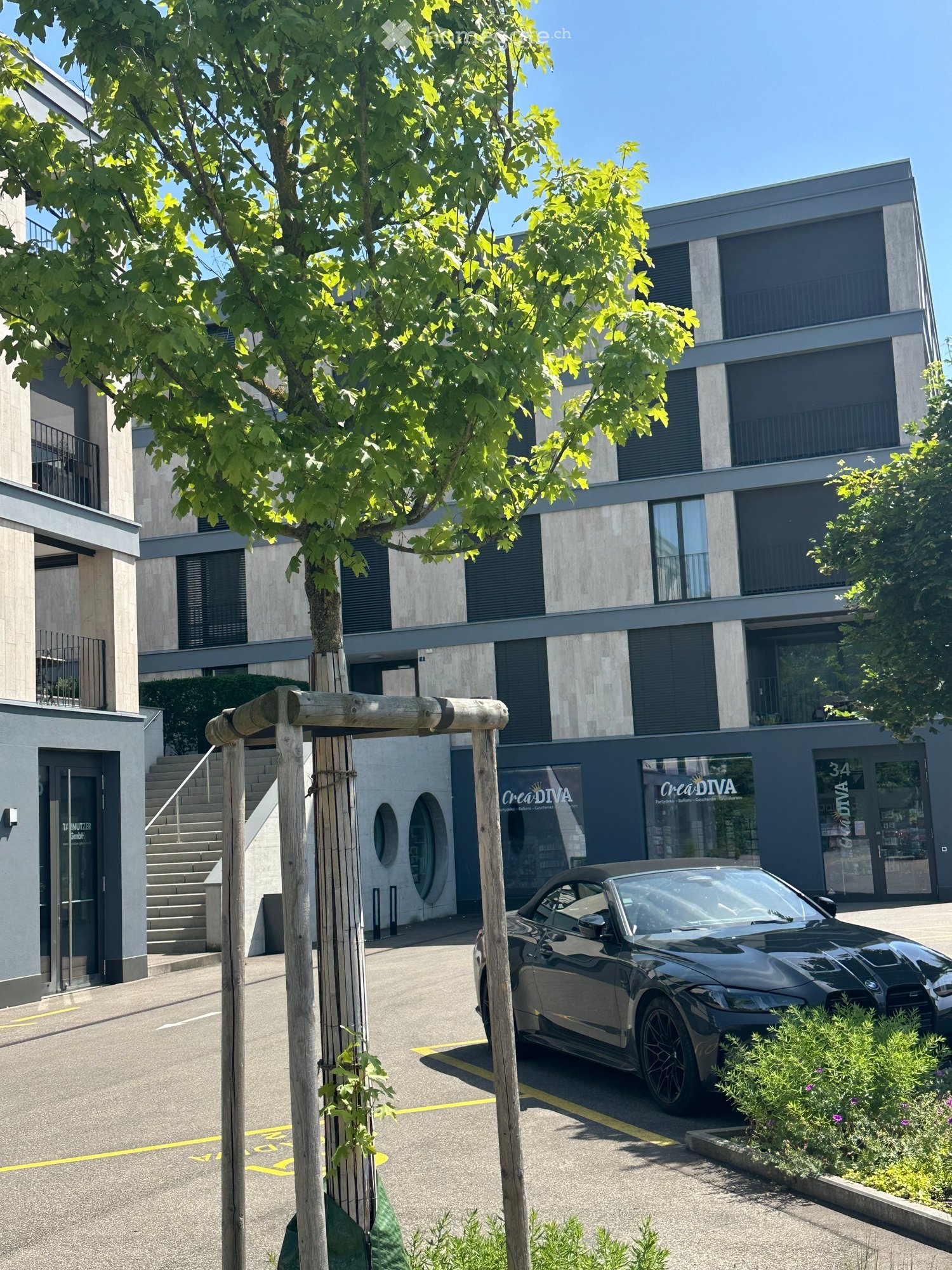 Modern multi-story building with large windows and balconies, featuring a sleek black convertible parked in front, surrounded by greenery and a wooden structure.