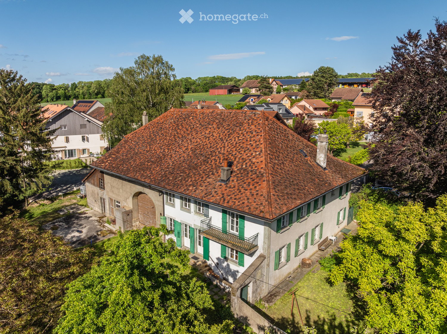 A two-story house with a red-tiled roof, white walls, green shutters, and a small balcony. The house is surrounded by greenery and trees, with neighboring houses visible in the background.