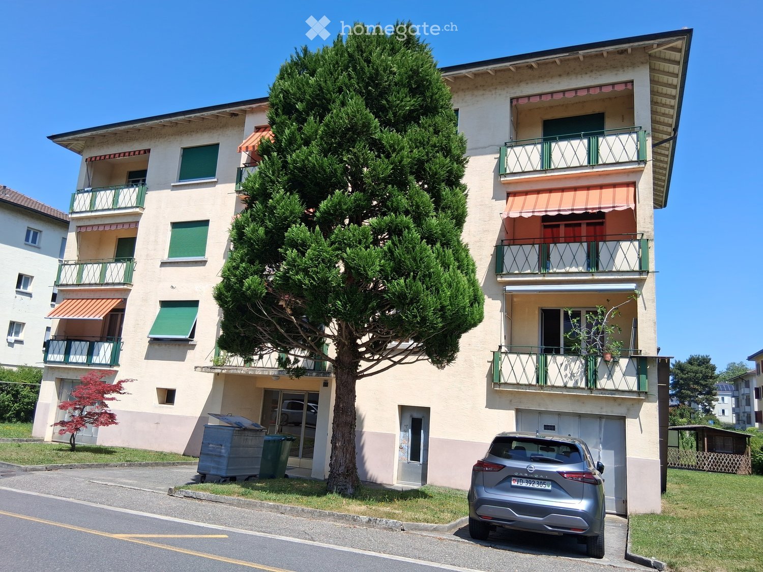 Four-story residential building with balconies, green shutters, and a large tree in front. A silver car is parked in front of the building.