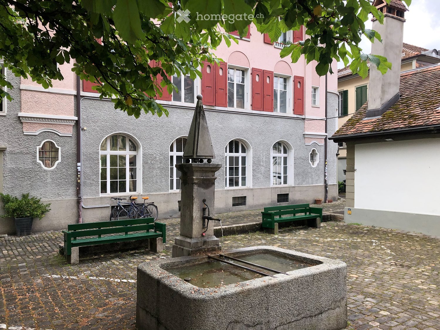Courtyard with a stone fountain, two green benches, and bicycles leaning against a building.