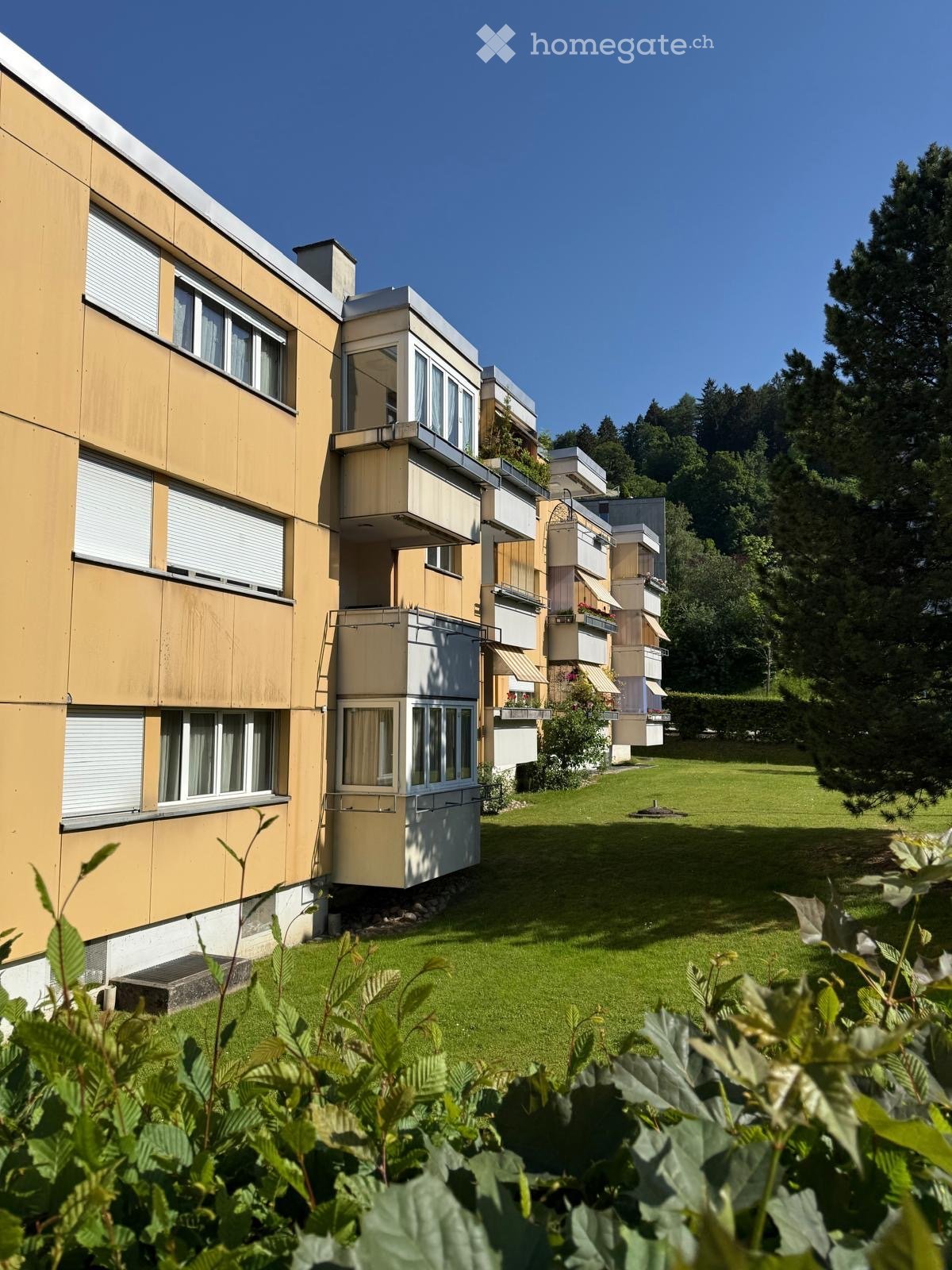 Modern multi-story residential building with balconies and large windows, surrounded by a well-maintained lawn and trees.