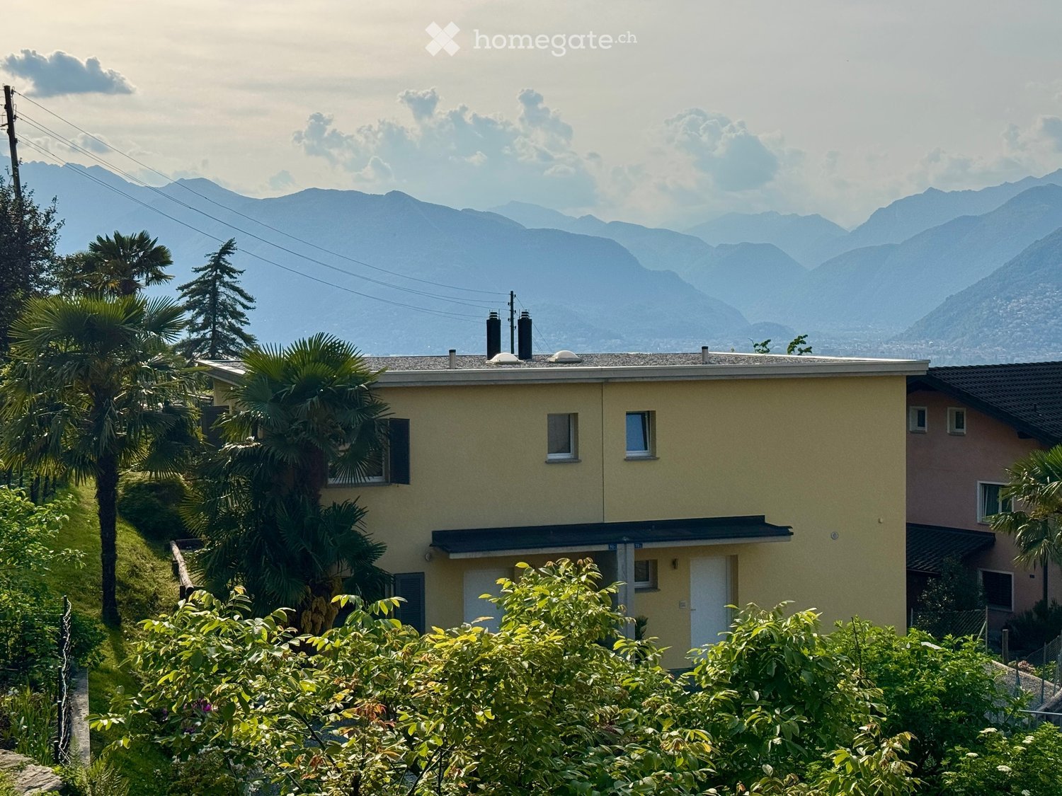 A two-story yellow house with white-framed windows and a gray roof, surrounded by lush greenery and palm trees, with a scenic mountain backdrop.