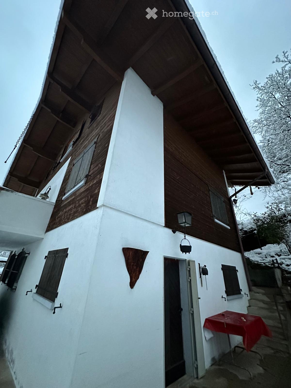 Two-story house with white walls, wooden trim, and a slanted roof. Features include wooden shutters, a small porch with a table covered in a red cloth, and a hanging lantern.