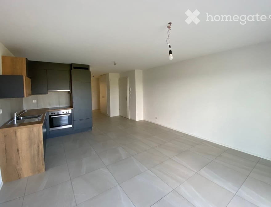 Empty room with tiled floor and white walls, connected to a modern kitchen area with wooden and grey cabinetry, stainless steel sink, and oven.