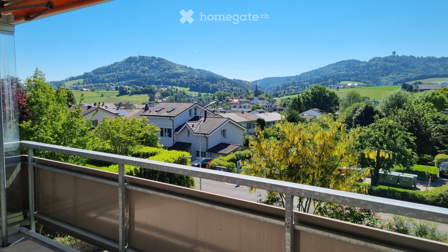 Balcony overlooking a scenic town with houses, trees, and distant hills under a clear blue sky.