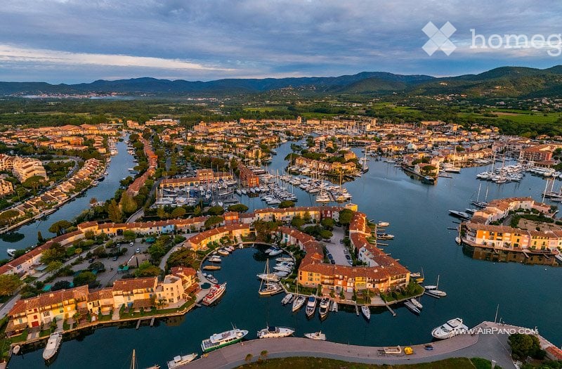 Aerial view of a coastal town with a marina filled with numerous boats and yachts, surrounded by colorful buildings and a scenic backdrop of hills and mountains.
