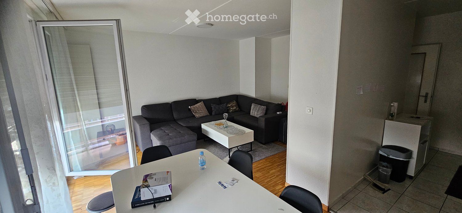 Modern living room with a grey sectional sofa, coffee table, dining table with chairs, and a balcony door with blinds.