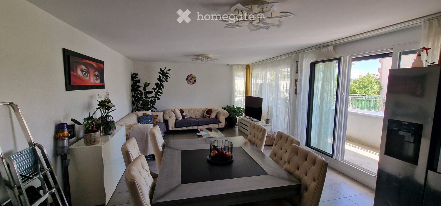 Bright living and dining area with a sofa, coffee table, dining table with chairs, and large windows leading to a balcony.