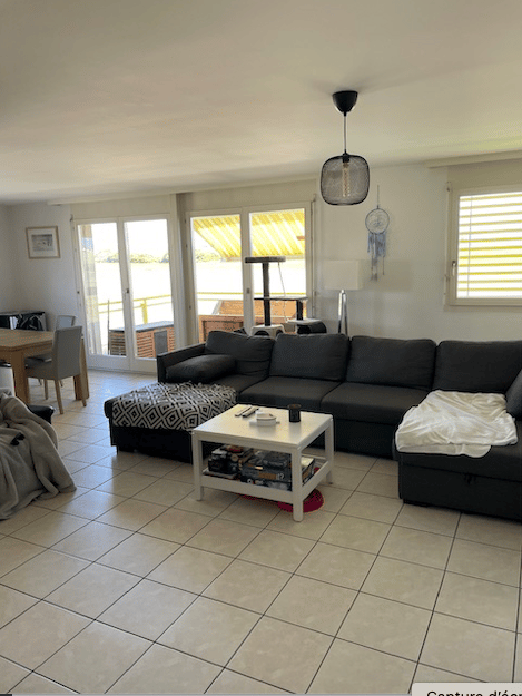 Living room with a grey sectional sofa, white coffee table, dining area with a table and chairs, large windows with blinds, and a view of a balcony area.