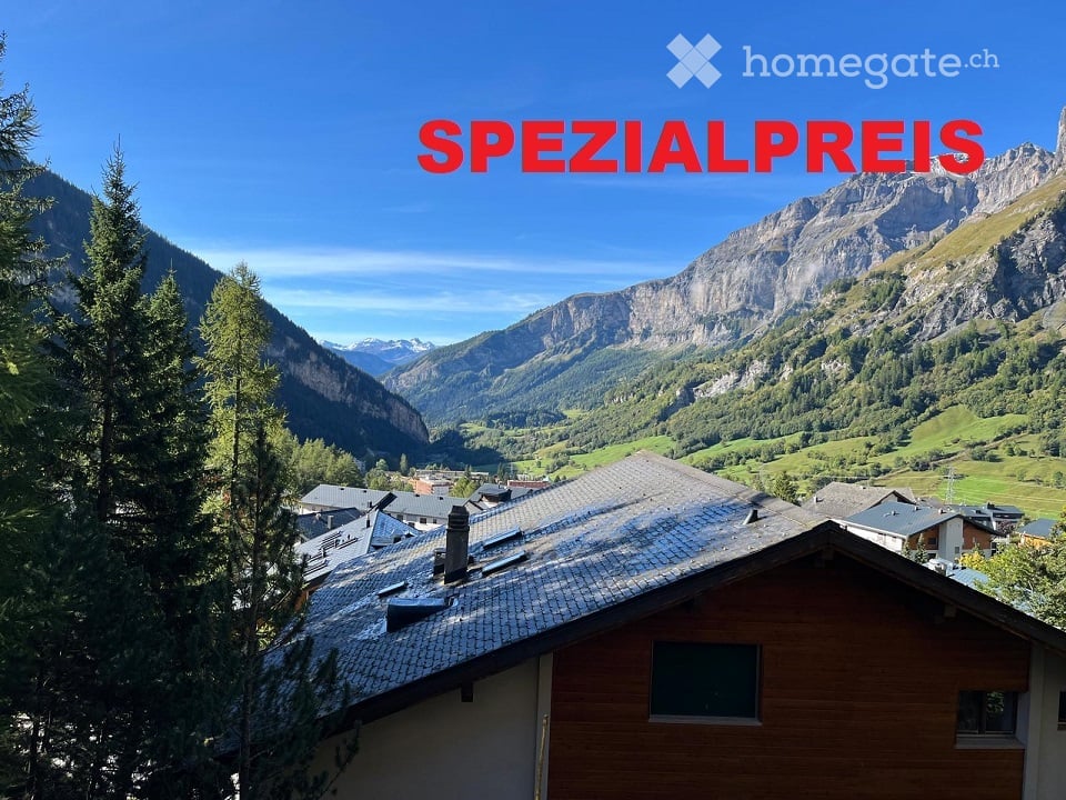 A wooden house with a tiled roof is situated in a picturesque valley surrounded by lush green trees and towering mountains under a clear blue sky.