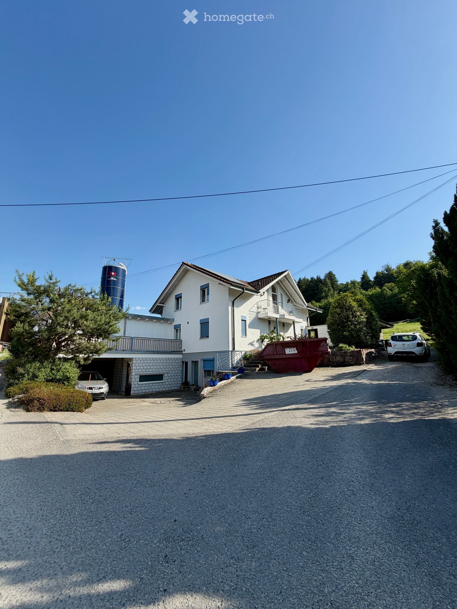 Two-story house with white exterior, multiple windows, garage, and balcony. There are two cars parked in the driveway and a silo in the background.