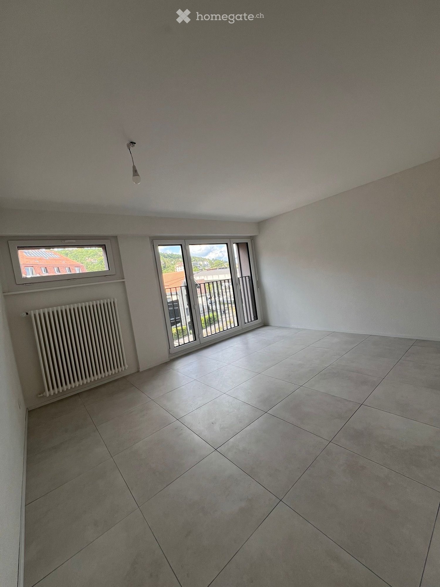 Empty room with tiled floor, white walls, radiator, large window, and sliding glass door leading to a balcony.