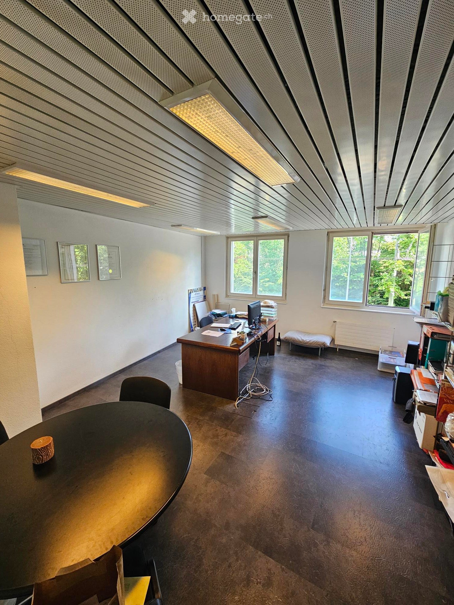 Office space with a large desk, computer, chairs, and a round table with a decorative item on it. The room has large windows with a view of greenery outside and a modern, minimalist design.