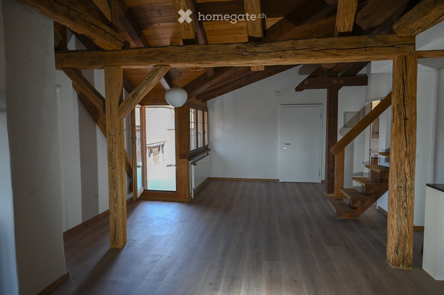 Spacious hallway with wooden beams and polished wooden flooring, leading to a staircase and an open doorway to an outdoor area.