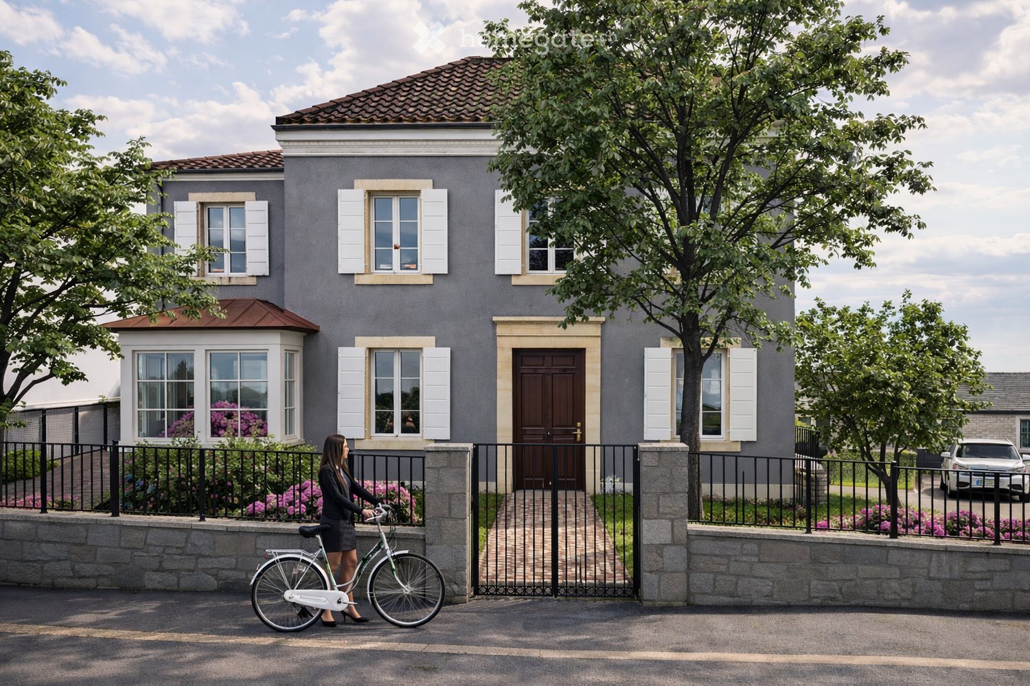 Two-story house with grey exterior, white shutters, and a brown front door. The house features a stone pathway leading to the entrance, surrounded by a black metal fence and flowering plants. A woman is riding a bicycle in front of the house.