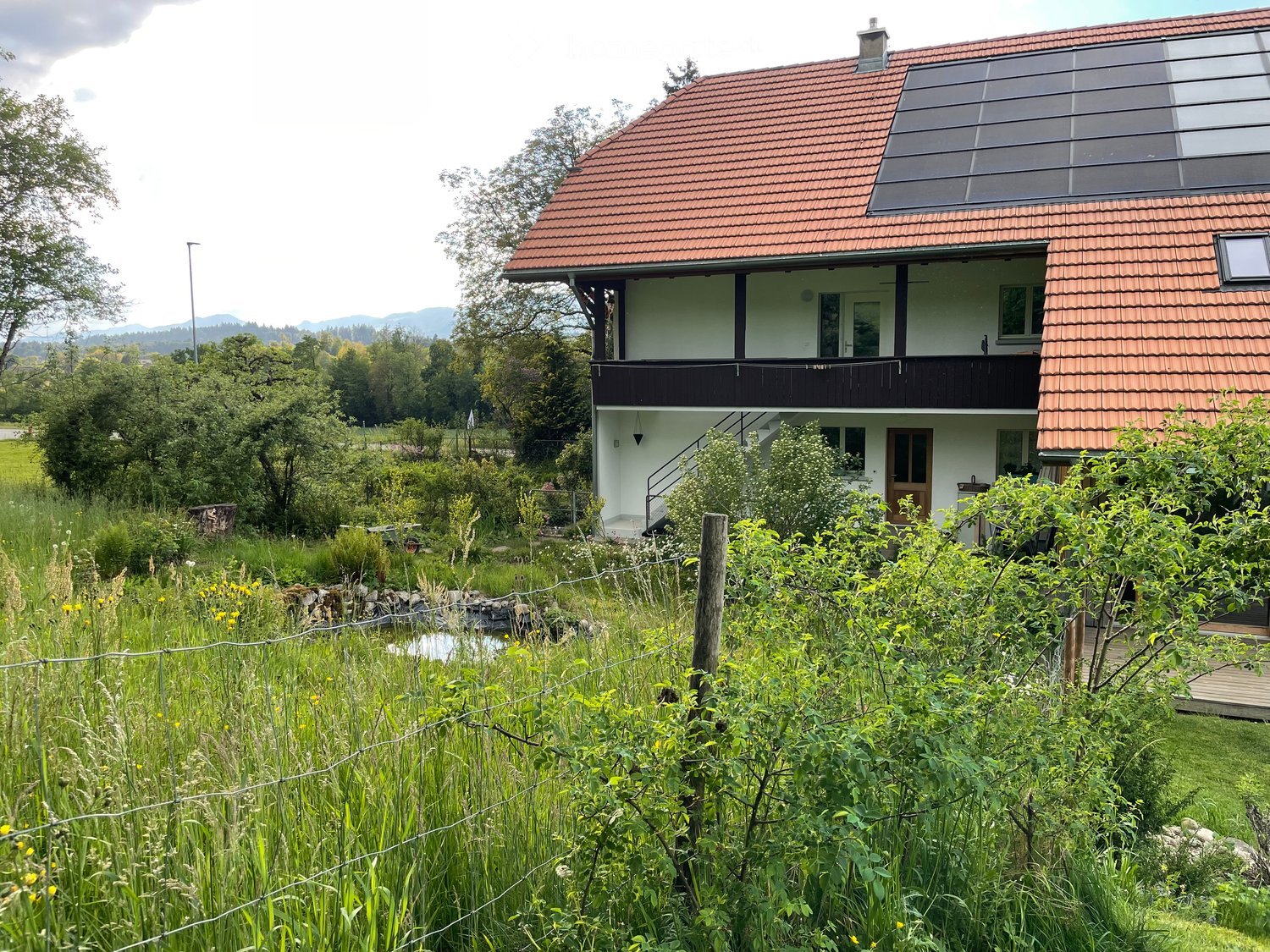 Two-story modern house with red-tiled roof, white walls, large windows, and a balcony overlooking a lush, green garden with a small pond and surrounding trees.
