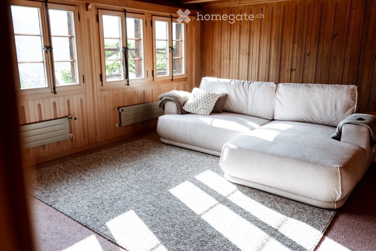 Cozy living room with wooden walls, large windows, a grey sectional sofa with pillows, and a grey rug on the floor.
