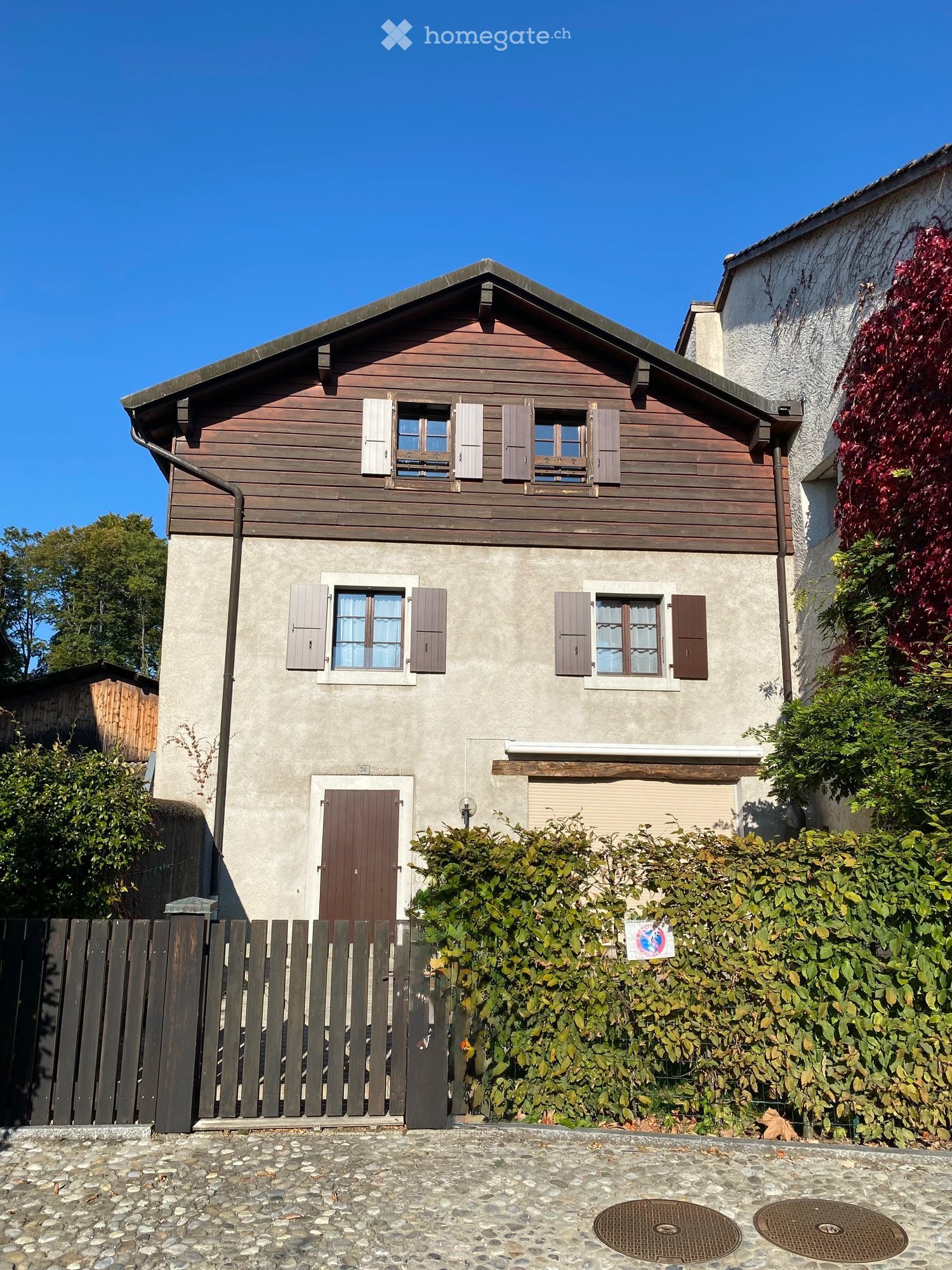 Two-story house with wooden siding on the upper level and concrete on the lower level, featuring multiple windows with shutters and a central front door.