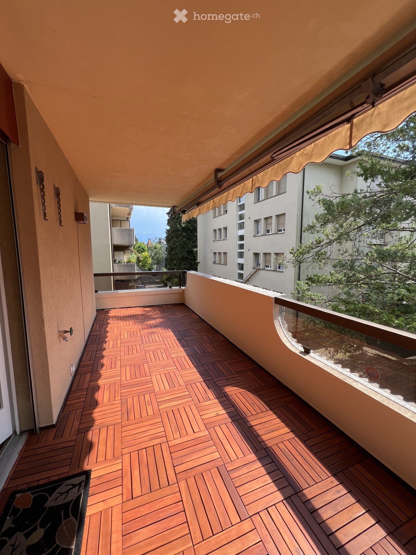 Wooden deck balcony with glass railing, beige awning, and view of neighboring buildings and greenery.