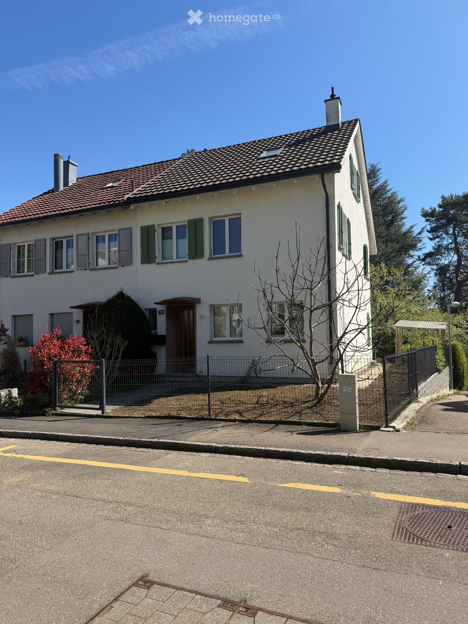 Two-story house with white walls, green shutters, red-tiled roof, and a fenced front yard with a small tree and flowering bush.
