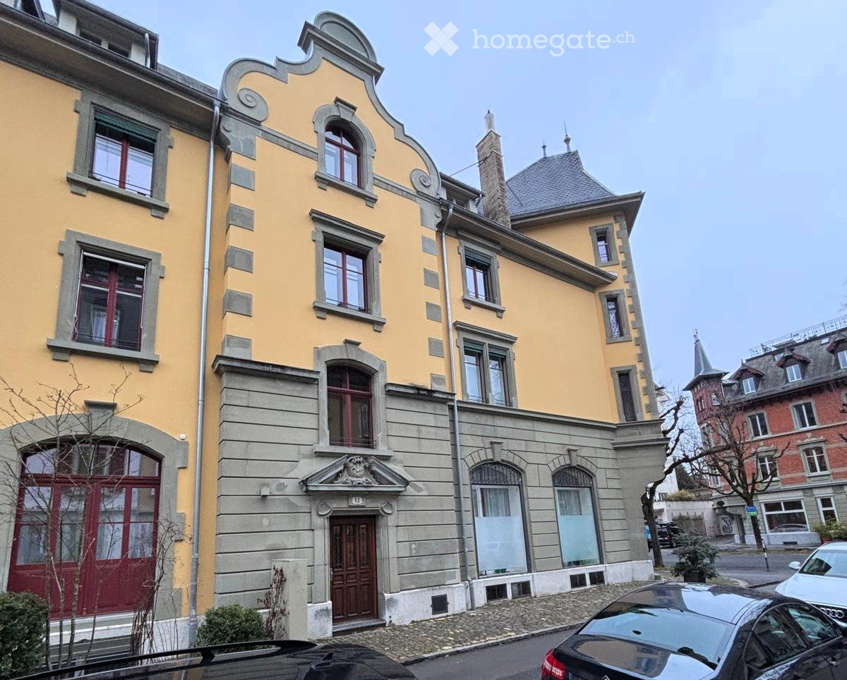 Building. A multi-story residential building with yellow facade, decorative stonework, multiple windows, and a chimney. The entrance features a wooden door with a decorative stone surround.