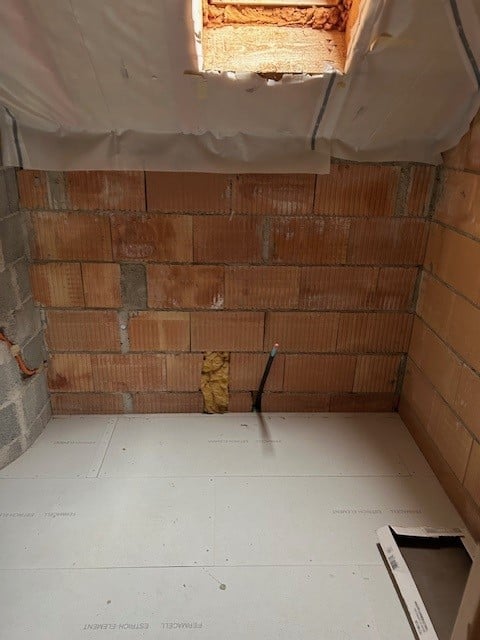 Empty bathroom under construction with exposed brick walls, white tile floor, and unfinished ceiling with insulation visible.