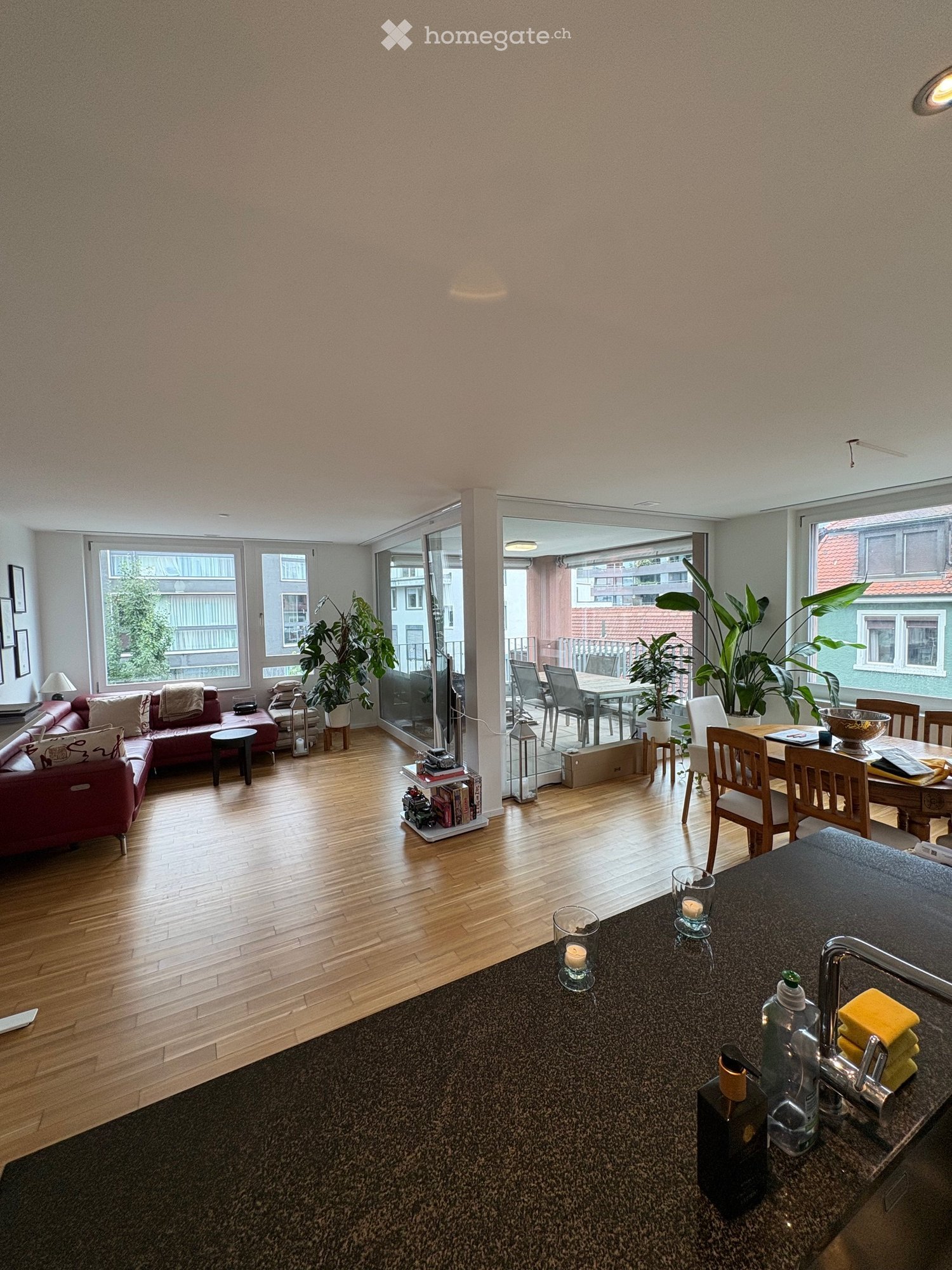 Open-plan living and dining area with wooden flooring, large windows, and a balcony. Features a red sofa, dining table with chairs, and a kitchen counter with a faucet and various items.