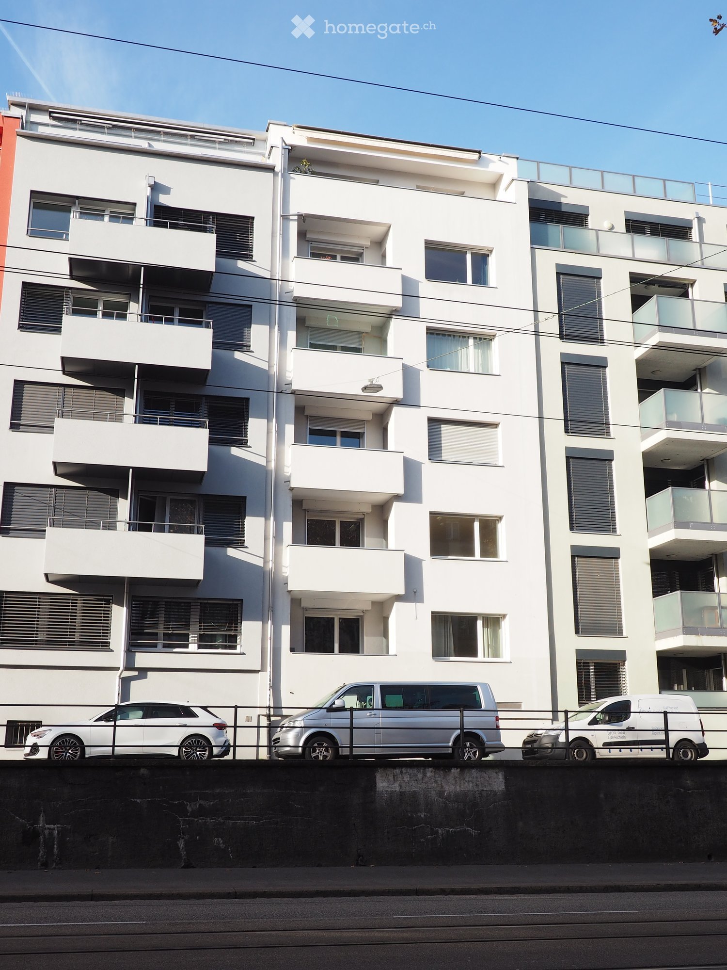 Modern multi-story residential building with white facade, multiple balconies, and large windows. Several cars are parked in front.