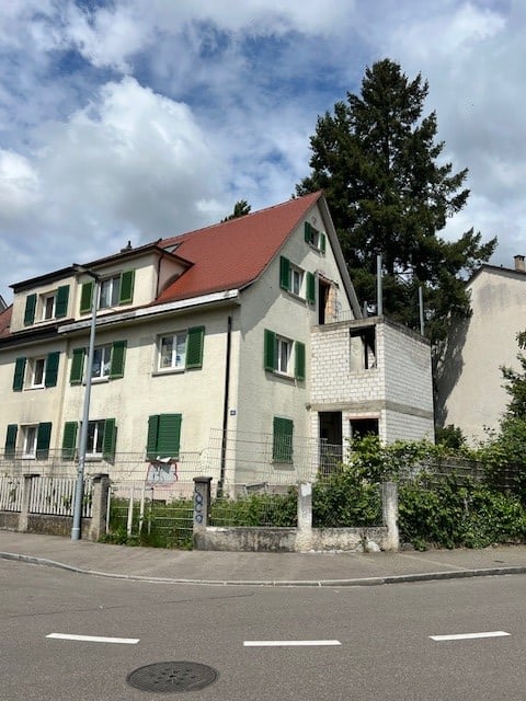 Two-story house with red-tiled roof, green shutters, and white walls. Features a small garden and fence in front.