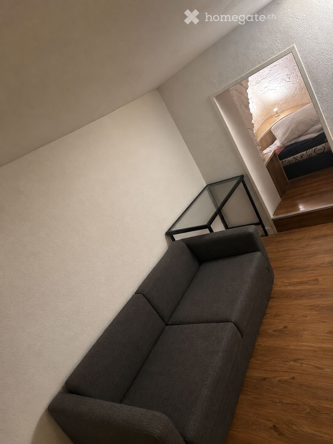 A modern living room with a grey sectional sofa, a sleek black glass table, and a large mirror reflecting a bedroom area with a bed and nightstand.