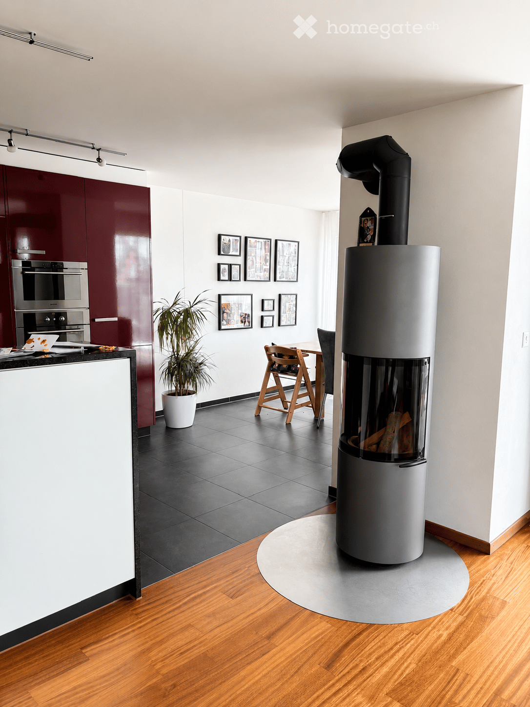Modern living space with a sleek, cylindrical wood-burning stove, a small dining area with a table and chairs, a potted plant, and a kitchen area with stainless steel appliances and dark red cabinets.