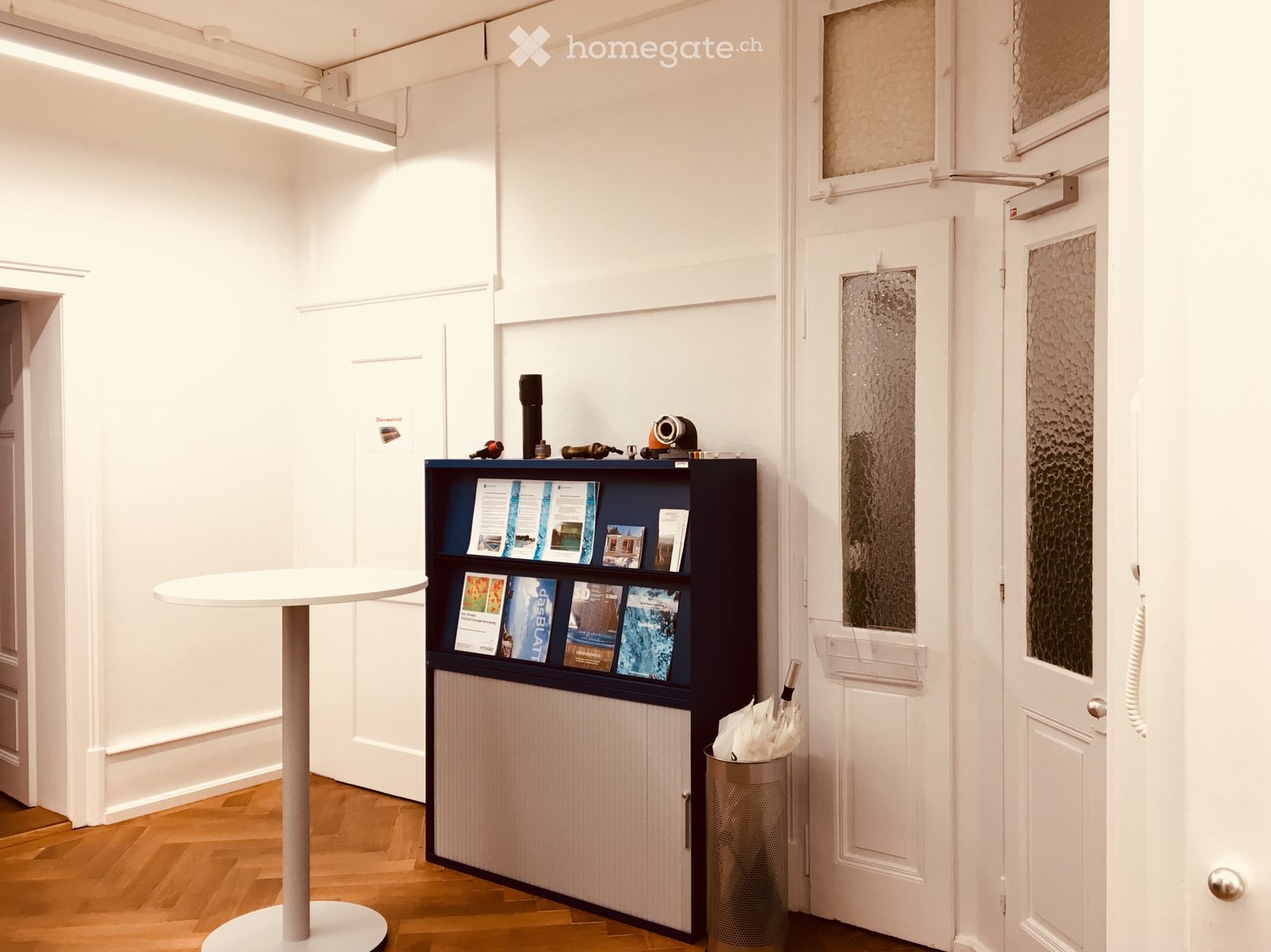 A modern, minimalistic hallway or reception area with a white table, a dark wooden shelf holding brochures, and a small umbrella stand. The floor is wooden, and the walls are white with framed artwork.