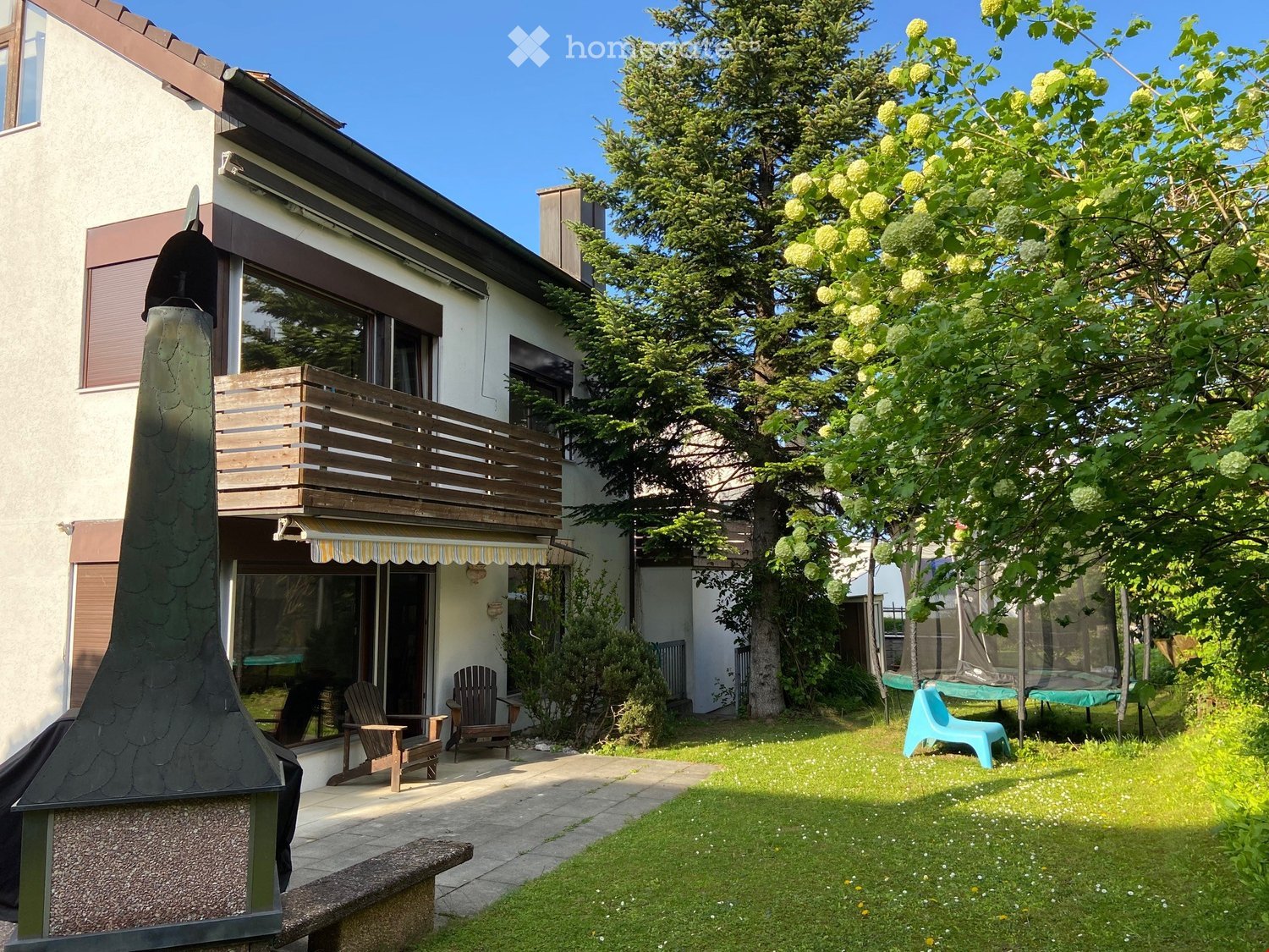 Two-story house with a balcony, wooden deck, and outdoor seating area. The garden includes a lawn, trees, and patio furniture.