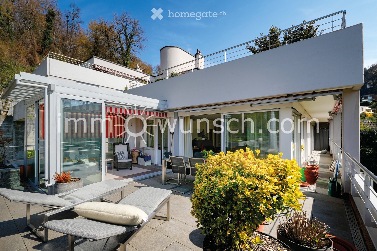 A modern rooftop terrace with lounge chairs, a dining table, potted plants, and a view of the surrounding greenery.