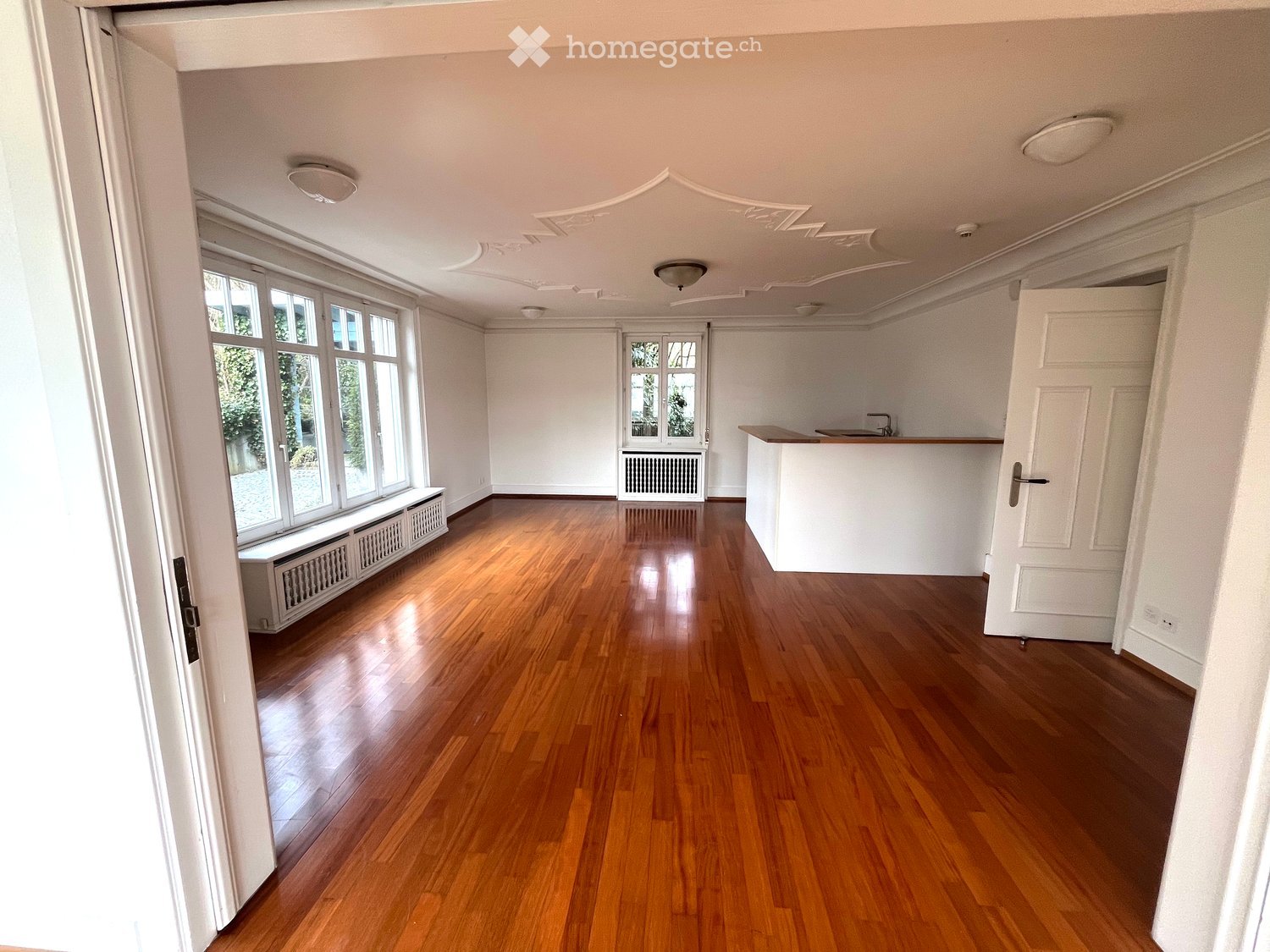 Empty room with wooden flooring, white walls, large windows, and a small counter area with a sink.