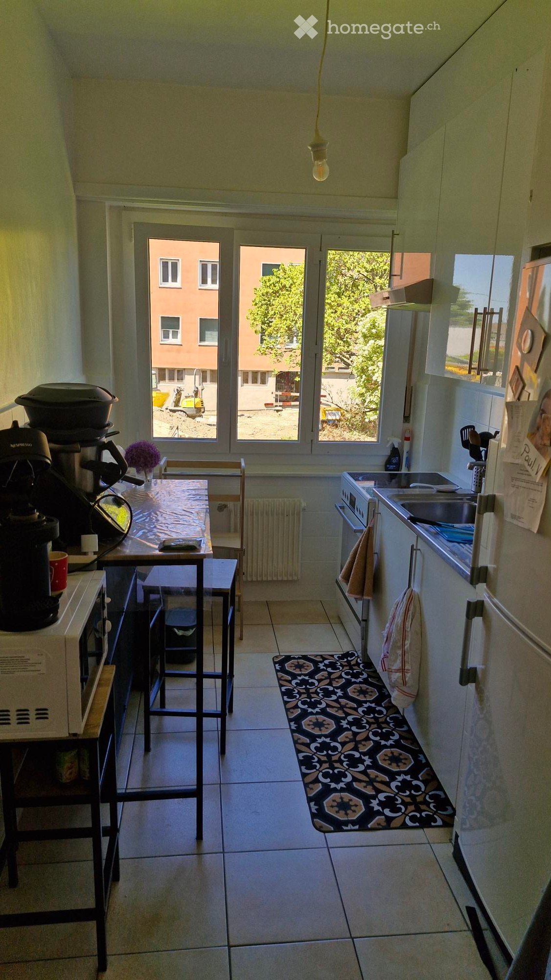 The kitchen features a coffee maker, a small table with a stool, an oven, a sink, and a refrigerator. There is a patterned rug on the floor and a flower vase on the table.