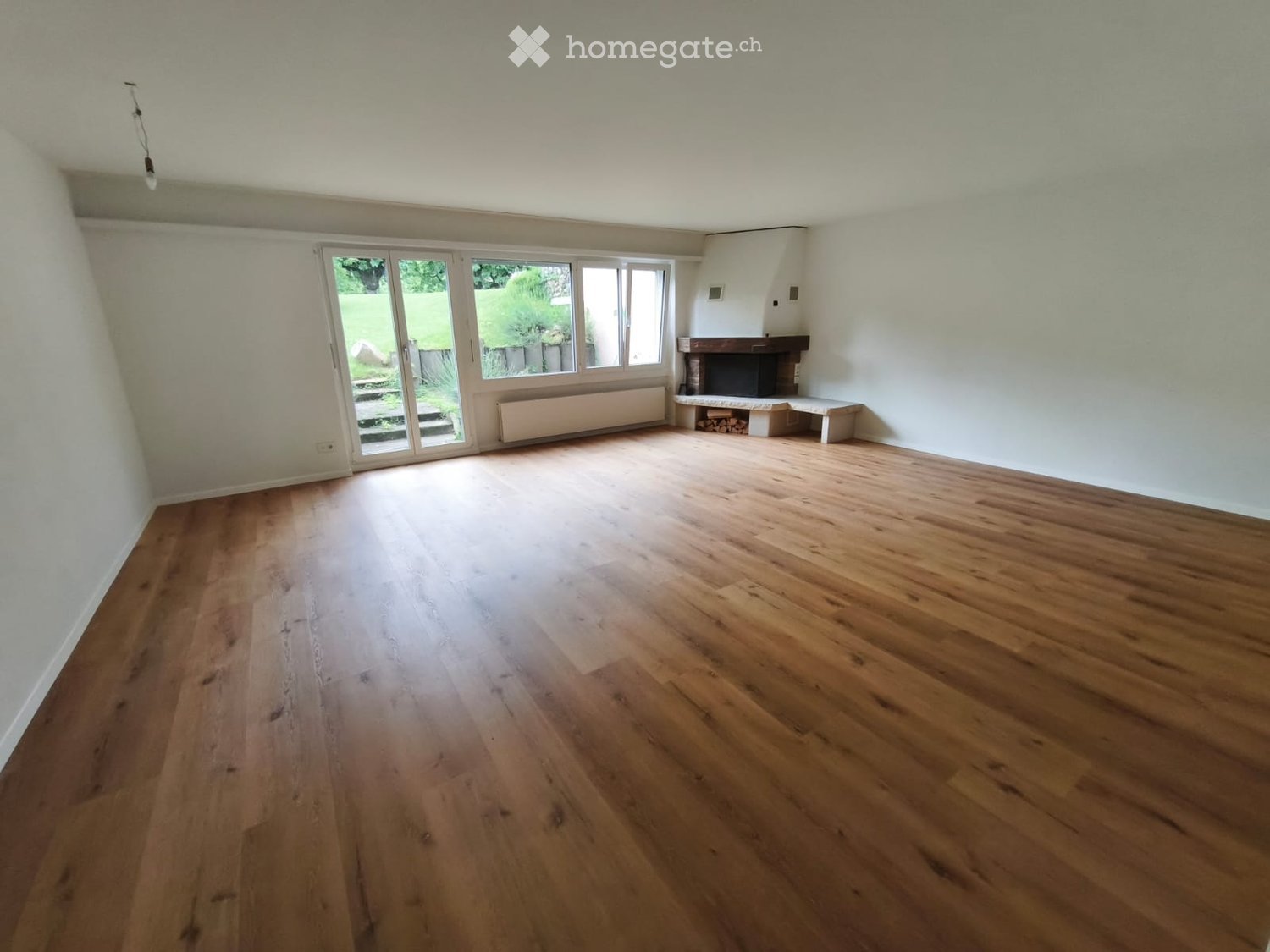 Empty room with wooden flooring, white walls, large sliding glass doors leading to a garden, and a fireplace with a bench.