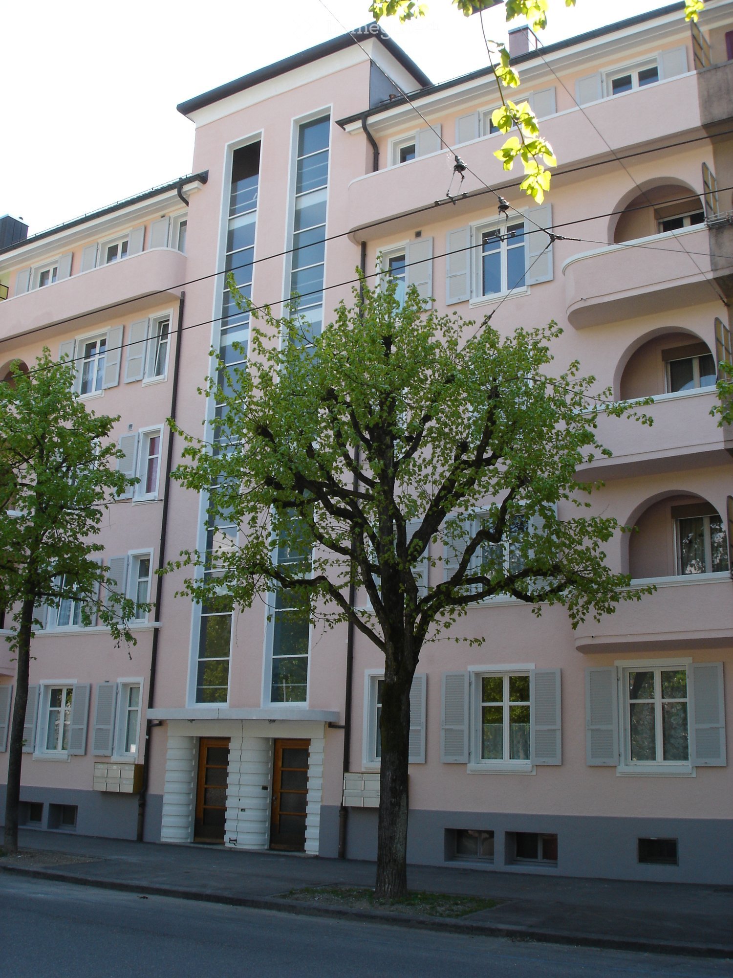 Pink multi-story building with white-framed windows, balconies, and a tree in front.