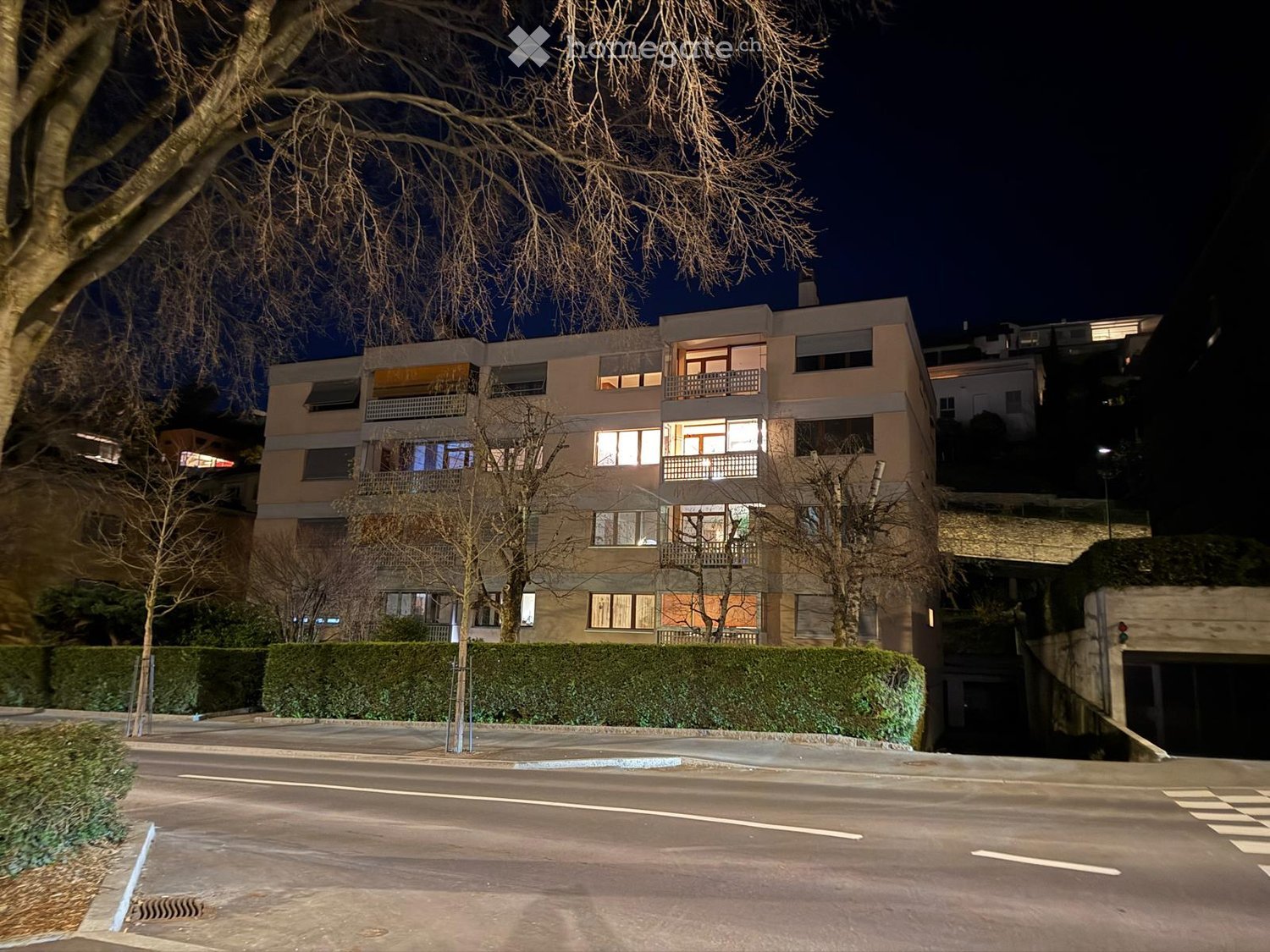 Multi-story residential building with illuminated windows and balconies, surrounded by trees and a hedge, with a street in front.