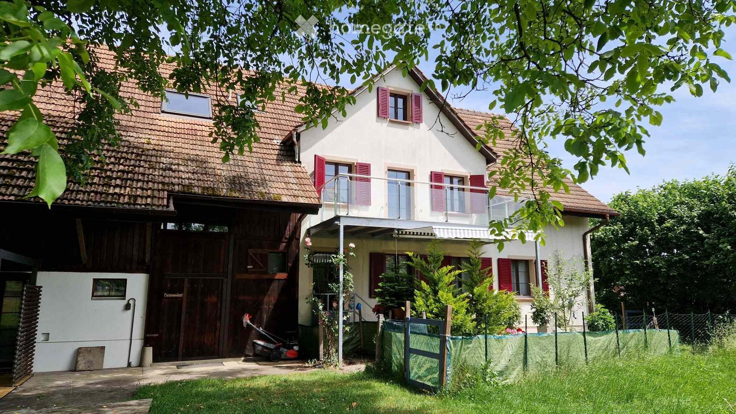 Two-story house with red shutters, tiled roof, balcony, and surrounding greenery.
