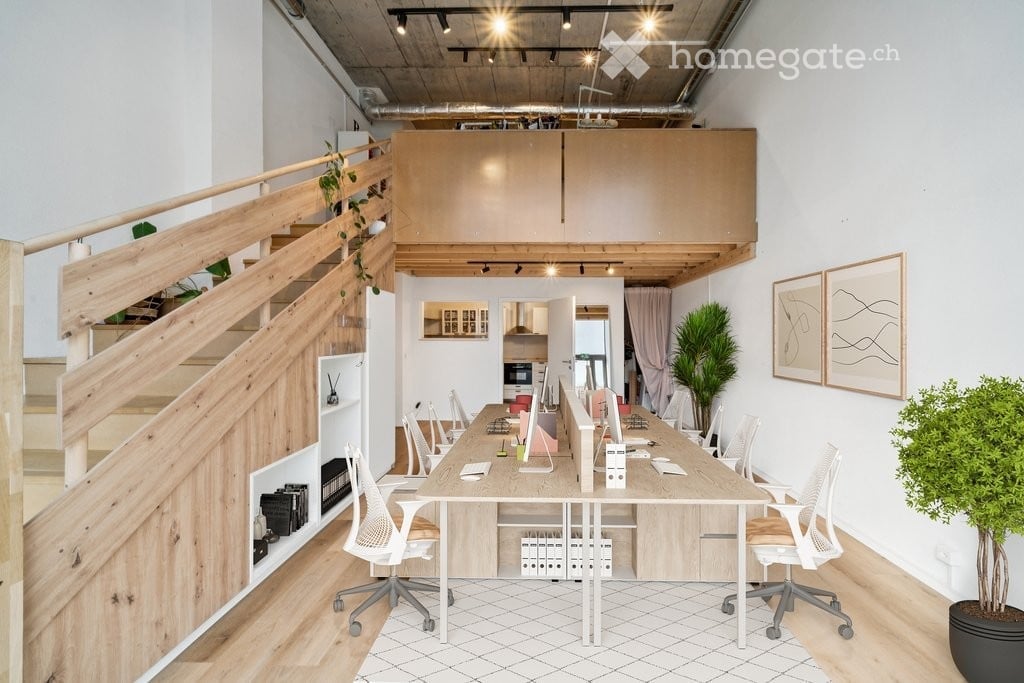 Modern office space with a long central table, ergonomic chairs, multiple screens, potted plants, and framed artwork on the walls. A wooden staircase leads to an upper level.