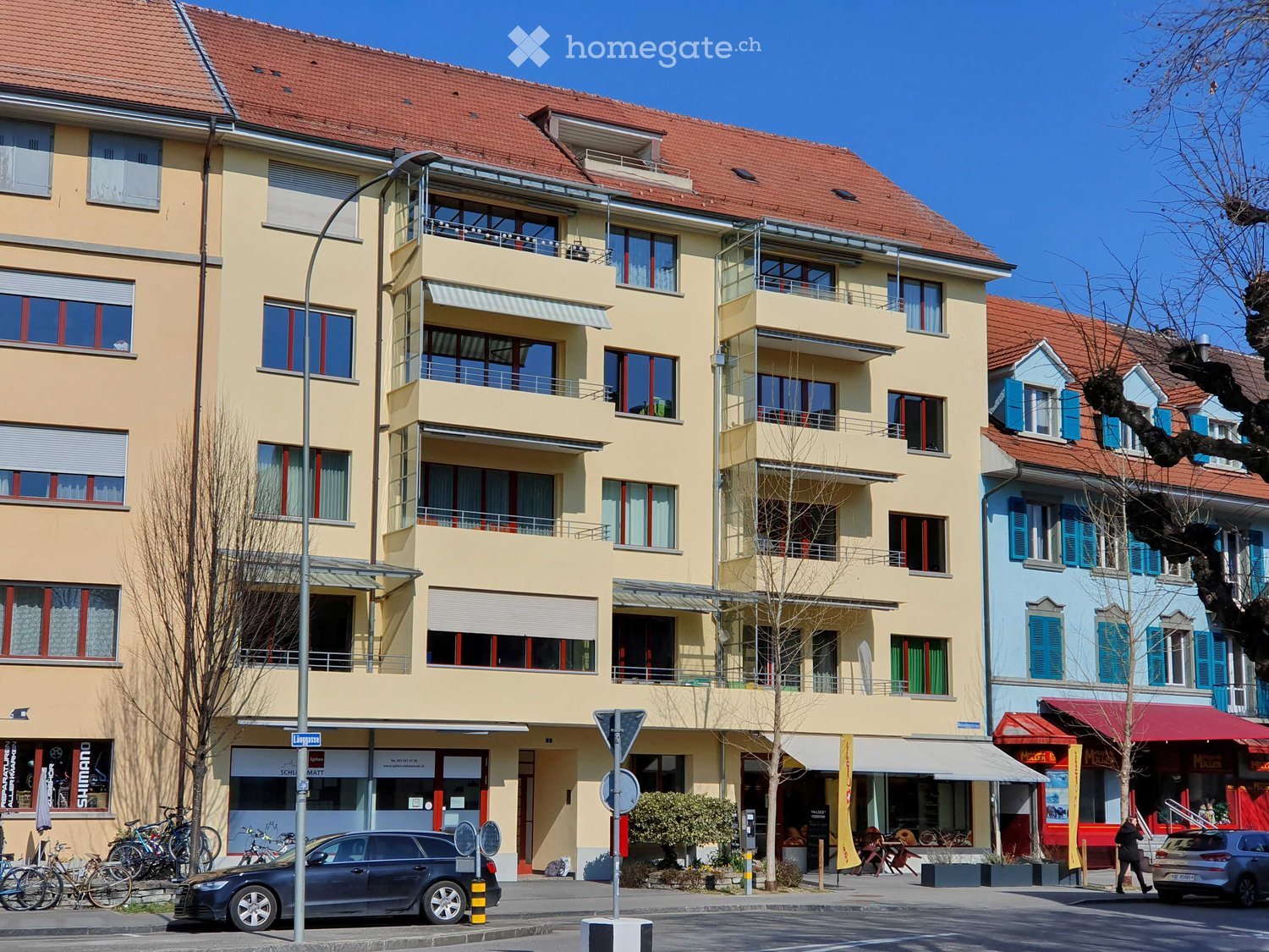 Multi-story residential building with balconies, large windows, and a red-tiled roof. The ground floor features commercial storefronts with awnings and signage. Bicycles and a car are parked on the street.